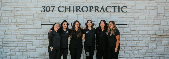 A close up of non-chiropractic staff standing outside in front of beige brick wall featuring the clinic name and logo.