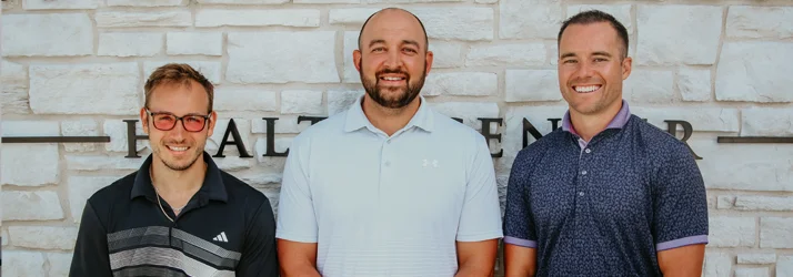 Medium shot of the three chiropractors at 307 Chiropractic standing against a beige brick wall outside.